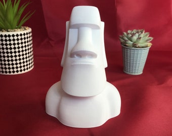 Könnte beinhalten: Eine weiße Moai-Statue, eine stilisierte Skulptur eines menschlichen Kopfes und Oberkörpers, steht auf einer roten Oberfläche. Die Statue hat eine große Nase und einen rechteckigen Kopfschmuck. Zwei kleine Topfpflanzen befinden sich auf beiden Seiten der Statue.