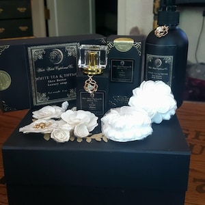 May include: A White Witch Cupboard Co. gift set featuring a black box, a black perfume bottle with a gold cap and floral charm, a black lotion pump bottle, and two white flower-shaped soaps. The set includes labels reading White Tea & Thyme.