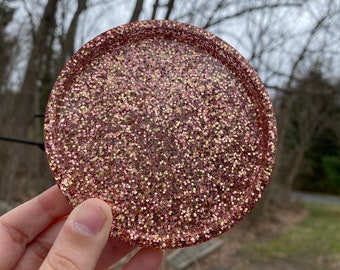 May include: A round, pink and gold glitter coaster. The coaster is made of resin and has a smooth, glossy finish.