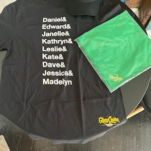 Può includere: Una maglietta nera con testo bianco che elenca i nomi, "Daniel, Edward, Janelle, Kathryn, Leslie, Kate, Dave, Jessica, Madelyn". La maglietta ha un logo giallo "Glass Onion" in basso a destra. Un cappello da baseball nero con il testo "A Rian Johnson Whodunnit" è sopra la maglietta. Un panno verde con il testo "Glass Onion" è sul lato destro della maglietta.