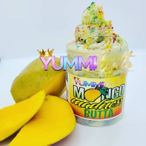 May include: A jar of mango-scented whipped body butter with a green and white swirl design. The jar has a label that says "YUMMI Butta" and "Mango Madness Butta". There are two slices of mango next to the jar.