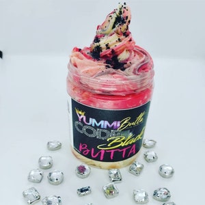 May include: A pink and white whipped body butter in a clear jar with a black label that reads "YUMMI Butta CODE Black BUTTA". The jar is surrounded by clear, faceted rhinestones.