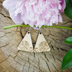 May include: A pair of wooden triangle earrings with a floral design. The earrings are light-colored wood with a laser-etched peony flower. The earrings hang from silver-colored hooks. A pink peony flower and green stem are in the background.