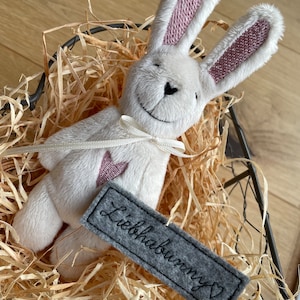 Könnte beinhalten: Ein weißer Plüschhase mit rosa Ohren und einem grauen Filzetikett mit der Aufschrift "Liebhabunny" mit einem Herz. Der Hase sitzt in einem Drahtkorb gefüllt mit zerkleinertem Papier.