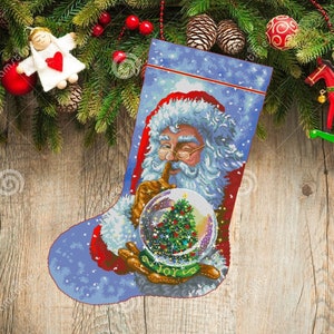 May include: A blue Christmas stocking featuring Santa Claus holding a snow globe with a Christmas tree inside. Santa has a finger to his lips, and the word "JOY" is at the bottom of the globe. The stocking is decorated with snowflakes.