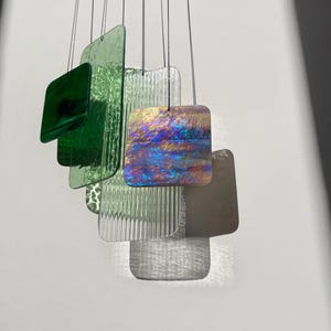 Op de afbeelding: Een hangende windgong met een messingkleurig bovenframe en meerdere glazen panelen. De panelen zijn in verschillende tinten groen, helder en iriserend, met vierkante en rechthoekige vormen. Het glas heeft een textuur en gladde oppervlakken.