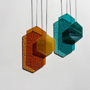 Könnte beinhalten: Abstrakte Hängekunst mit geometrischen Formen in Orange, Gelb und Türkis. Die Formen sind an dünnen schwarzen Schnüren aufgehängt und erzeugen eine moderne Darstellung. Das Glas hat eine strukturierte Oberfläche. Maße in Zentimetern.