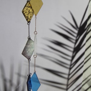 K&ouml;nnte beinhalten: Ein H&auml;ngemobil mit drei diamantf&ouml;rmigen Glasst&uuml;cken in Gold, Wei&szlig; und Blau, mit einem Kristallprisma am Boden.