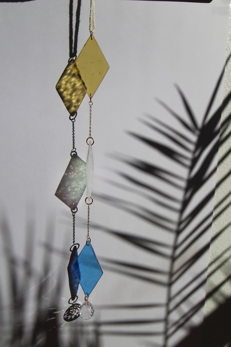 K&ouml;nnte beinhalten: Ein H&auml;ngemobil mit drei diamantf&ouml;rmigen Glasst&uuml;cken in Gold, Wei&szlig; und Blau, mit einem Kristallprisma am Boden.