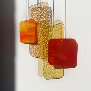 Op de afbeelding: Een hangende windgong met rechthoekige en vierkante glazen stukken in tinten oranje, goud en rood. Het glas heeft een getextureerd en glad oppervlak, opgehangen aan zwarte touwtjes. Het ontwerp is modern en decoratief.