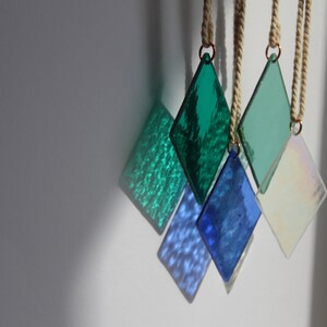 K&ouml;nnte beinhalten: F&uuml;nf diamantf&ouml;rmige Glasornamente, die an Bindf&auml;den h&auml;ngen. Die Ornamente sind in Gr&uuml;n- und Blaut&ouml;nen gehalten, wobei ein Ornament irisierend ist.