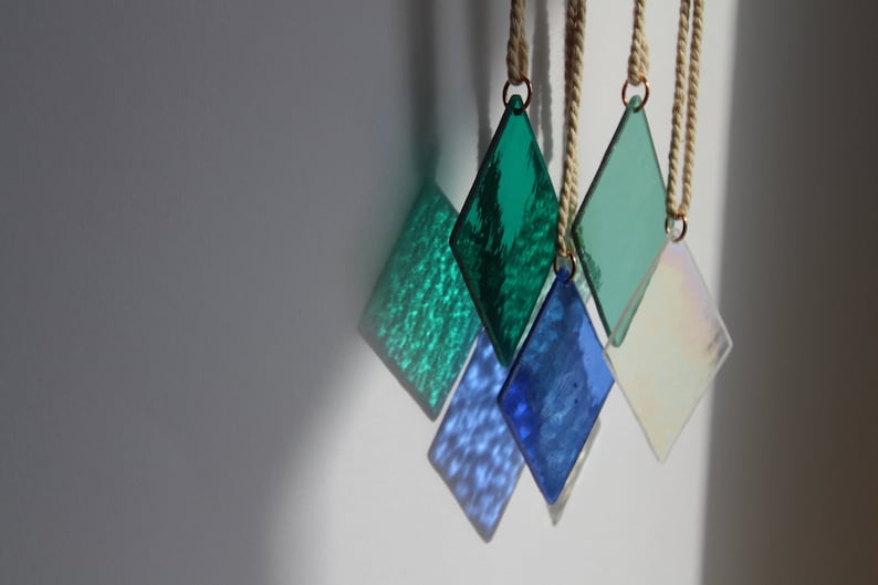 K&ouml;nnte beinhalten: F&uuml;nf diamantf&ouml;rmige Glasornamente, die an Bindf&auml;den h&auml;ngen. Die Ornamente sind in Gr&uuml;n- und Blaut&ouml;nen gehalten, wobei ein Ornament irisierend ist.