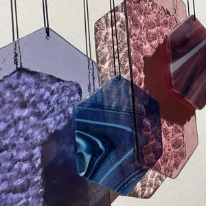 Può includere: Cinque ornamenti in vetro esagonali appesi a delle corde. Gli ornamenti sono fatti di diversi tipi di vetro, tra cui vetro trasparente, blu e viola. Gli ornamenti sono tutti di dimensioni e forme diverse.