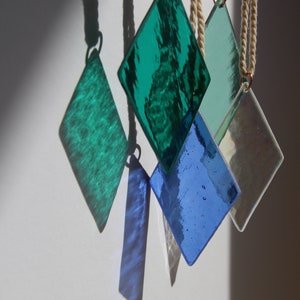 K&ouml;nnte beinhalten: F&uuml;nf diamantf&ouml;rmige Glasornamente, die an wei&szlig;en Schn&uuml;ren h&auml;ngen. Die Ornamente sind gr&uuml;n, blau und schillernd.