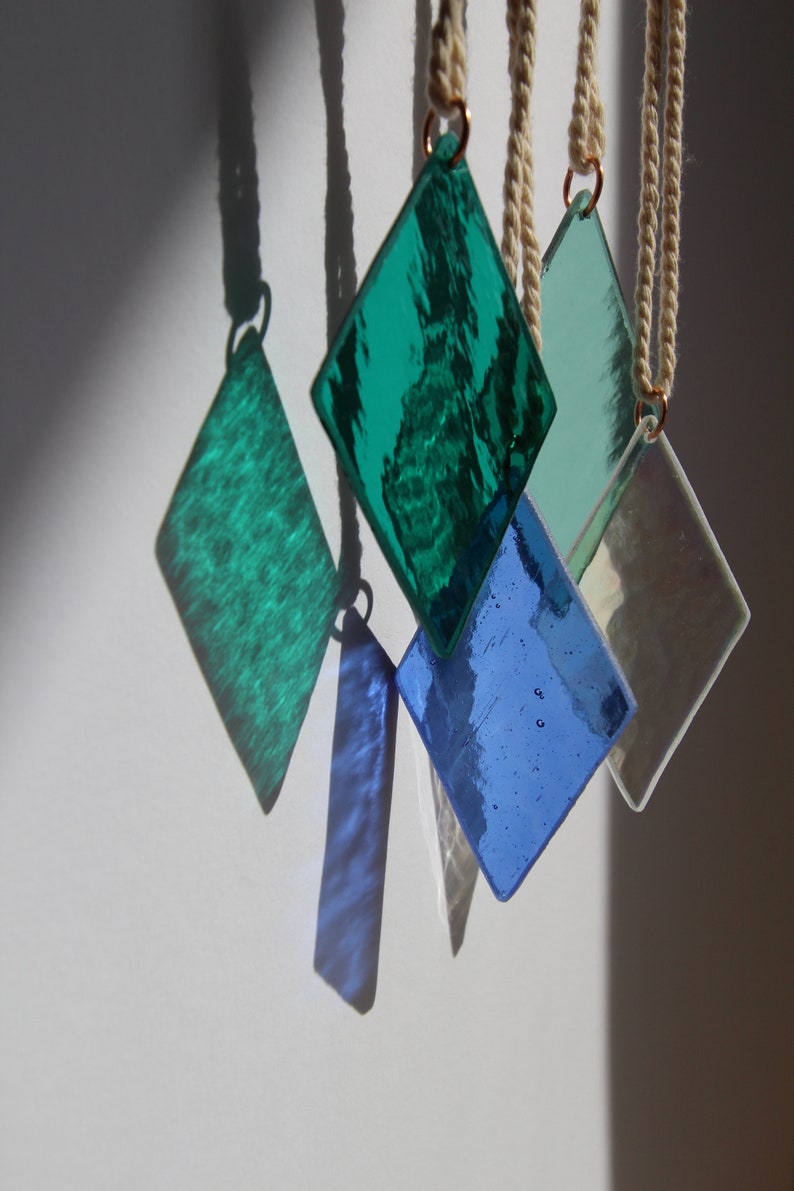 K&ouml;nnte beinhalten: F&uuml;nf diamantf&ouml;rmige Glasornamente, die an wei&szlig;en Schn&uuml;ren h&auml;ngen. Die Ornamente sind gr&uuml;n, blau und schillernd.