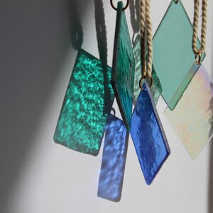 K&ouml;nnte beinhalten: Eine Sammlung von sechs Glasornamenten, jedes mit einer anderen Farbe und Textur. Die Ornamente h&auml;ngen an wei&szlig;en Schn&uuml;ren von einem wei&szlig;en Hintergrund.
