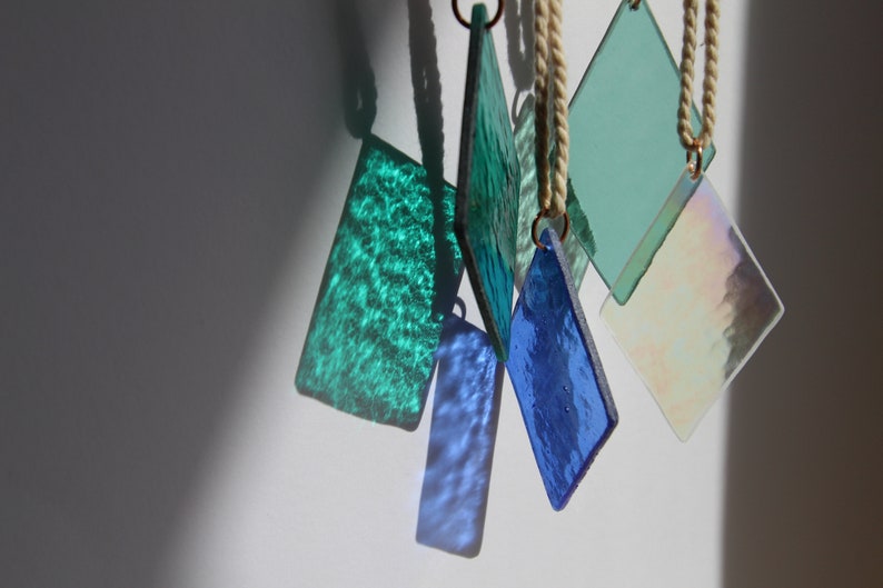 K&ouml;nnte beinhalten: Eine Sammlung von sechs Glasornamenten, jedes mit einer anderen Farbe und Textur. Die Ornamente h&auml;ngen an wei&szlig;en Schn&uuml;ren von einem wei&szlig;en Hintergrund.