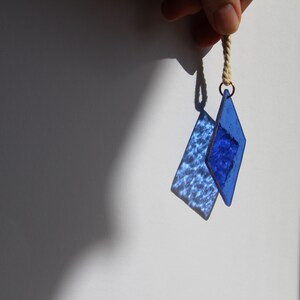 K&ouml;nnte beinhalten: Zwei blaue, diamantf&ouml;rmige Glasornamente, die an einer wei&szlig;en Schnur h&auml;ngen. Die Ornamente sind strukturiert und haben einen hellblauen Farbton.
