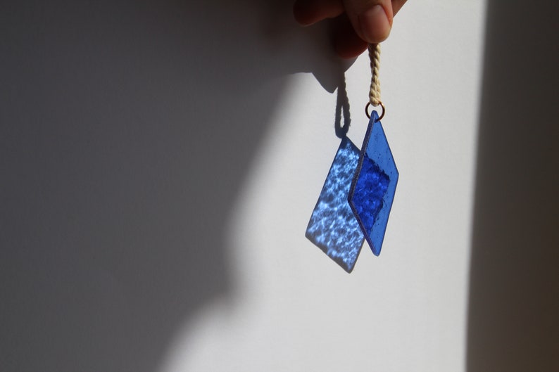 K&ouml;nnte beinhalten: Zwei blaue, diamantf&ouml;rmige Glasornamente, die an einer wei&szlig;en Schnur h&auml;ngen. Die Ornamente sind strukturiert und haben einen hellblauen Farbton.