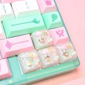 May include: A close-up of a pastel-colored keyboard with custom keycaps. The keys are white, mint green, and pink, with various symbols and designs. Some keycaps feature miniature cupcake and heart designs. The keyboard has a mint green frame.
