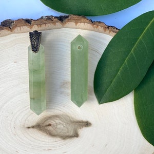 Puede incluir: Dos colgantes de cristal verde con un cierre de metal dorado. Los colgantes est&aacute;n sobre una superficie de madera con hojas verdes.