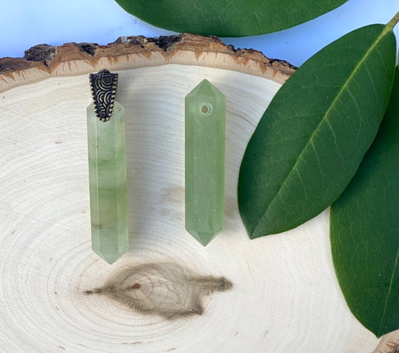 Puede incluir: Dos colgantes de cristal verde con un cierre de metal dorado. Los colgantes est&aacute;n sobre una superficie de madera con hojas verdes.