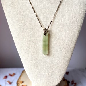 Puede incluir: Un collar con un colgante de cristal verde en una cadena de bronce con un cierre adornado. El colgante es un cristal largo y puntiagudo de color verde claro.