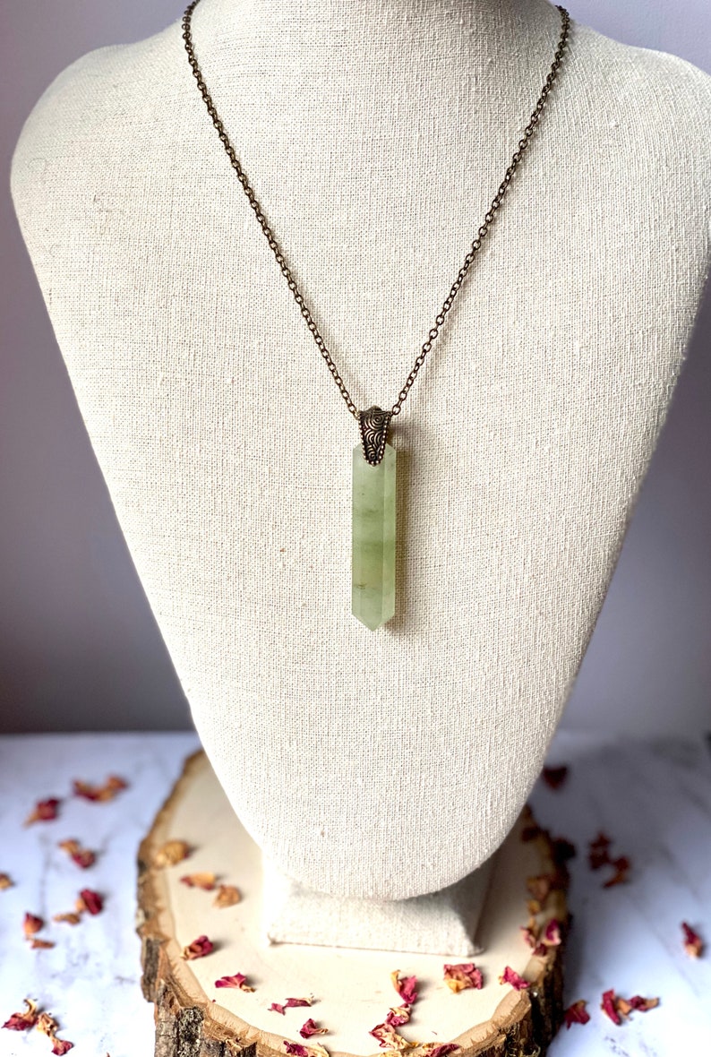 Puede incluir: Un collar con un colgante de cristal verde en una cadena de bronce con un cierre adornado. El colgante es un cristal largo y puntiagudo de color verde claro.