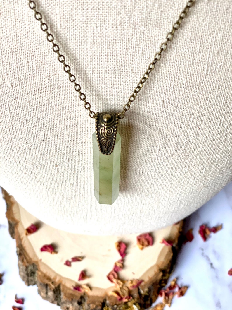 Puede incluir: Un collar con un colgante de cristal verde, una cadena de lat&oacute;n y un cierre de lat&oacute;n adornado. El colgante es un cristal largo y puntiagudo con un dise&ntilde;o facetado.