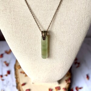 Puede incluir: Un collar con un colgante de cristal verde, una cadena de lat&oacute;n y un cierre adornado. El colgante tiene una forma rectangular alargada con una superficie lisa y pulida.