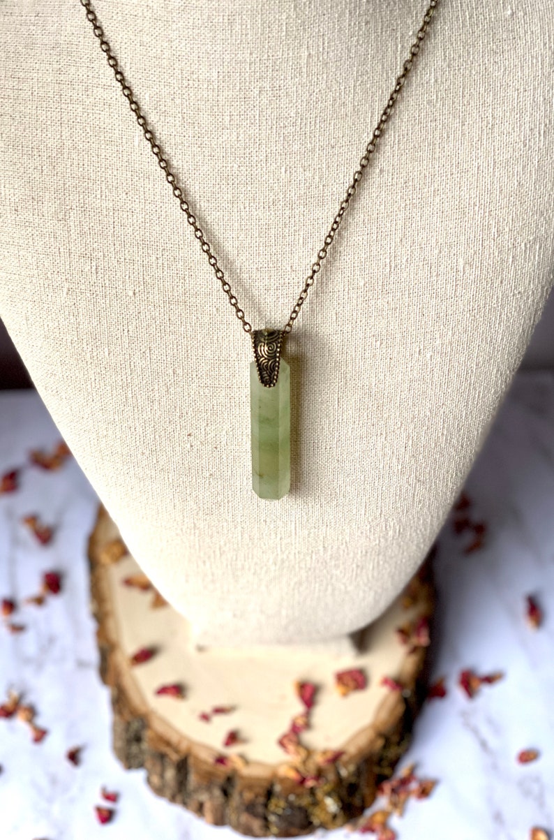 Puede incluir: Un collar con un colgante de cristal verde, una cadena de lat&oacute;n y un cierre adornado. El colgante tiene una forma rectangular alargada con una superficie lisa y pulida.