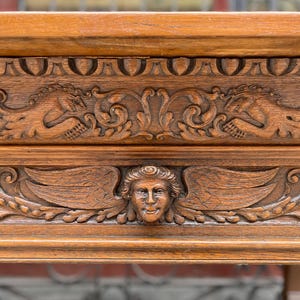 Może przedstawiać: Zbliżenie na rzeźbioną drewnianą kominkową półkę z misternymi detalami. Półka kominkowa ma centralną twarz cheruba z skrzydłami i dwie boczne figury z skrzydłami.