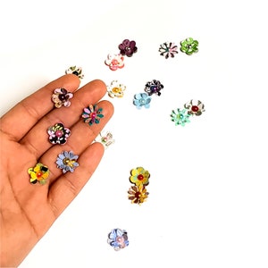 May include: Assortment of colorful paper flowers with a pearl center. The flowers are arranged on a white surface and a hand is holding some of the flowers.