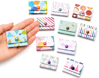 May include: A collection of ten small, rectangular paper envelopes featuring various colorful patterns including balloons, sprinkles, donuts, stripes, confetti, candles, macarons, and birthday text, each secured with a single round bead.
