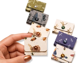May include: A collection of small, rectangular paper envelopes featuring various insect patterns, including dragonflies and beetles. Each envelope is secured with a single, centered, round bead acting as a button closure in metallic or pearl finishes.