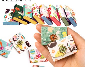 May include: A collection of miniature, patterned boxes and small, floral-adorned pouches. The boxes feature colorful designs with bunnies, flowers, and Easter eggs. The pouches are held in a hand, showcasing a gold-colored flower clasp.