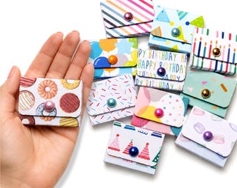May include: A collection of small, rectangular paper envelopes featuring various colorful patterns including donuts, birthday candles, party hats, and confetti. Each envelope is secured with a single, round, metallic-finish bead button.