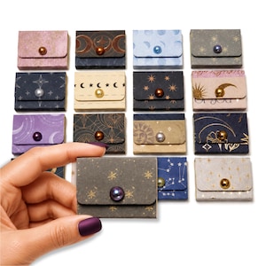 May include: A collection of small, rectangular paper envelopes featuring various celestial patterns, including moons, stars, and suns. Each envelope is secured with a single, centered, round bead in colors like gold, silver, purple, and bronze.