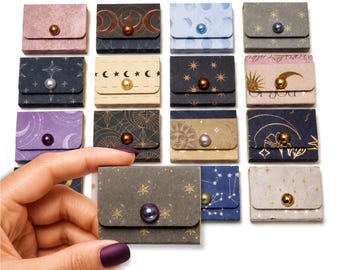 May include: A collection of small, rectangular paper envelopes featuring various celestial patterns, including moons, stars, and suns. Each envelope is secured with a single, centered, round bead in colors like gold, silver, purple, and bronze.