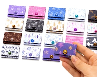 May include: A collection of small, rectangular, folded paper pouches featuring various holiday-themed patterns including snowflakes, trees, and skates. Each pouch is secured with a single, colorful, round bead acting as a button closure.