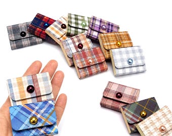 May include: A collection of small, rectangular, plaid-patterned fabric pouches, each featuring a single decorative bead closure on the front flap. The pouches come in various color combinations including blue, red, green, and tan. Text reads Includes One.