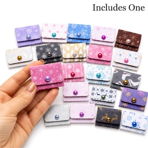 May include: A collection of small, rectangular, folded paper envelopes featuring various winter-themed patterns, including snowflakes, reindeer, and trees. Each envelope is secured with a single, colorful, round bead acting as a button closure.