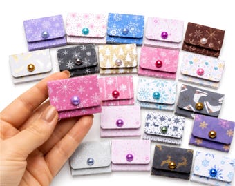 May include: A collection of small, rectangular, folded paper envelopes featuring various winter-themed patterns, including snowflakes, reindeer, and trees. Each envelope is secured with a single, colorful, round bead acting as a button closure.