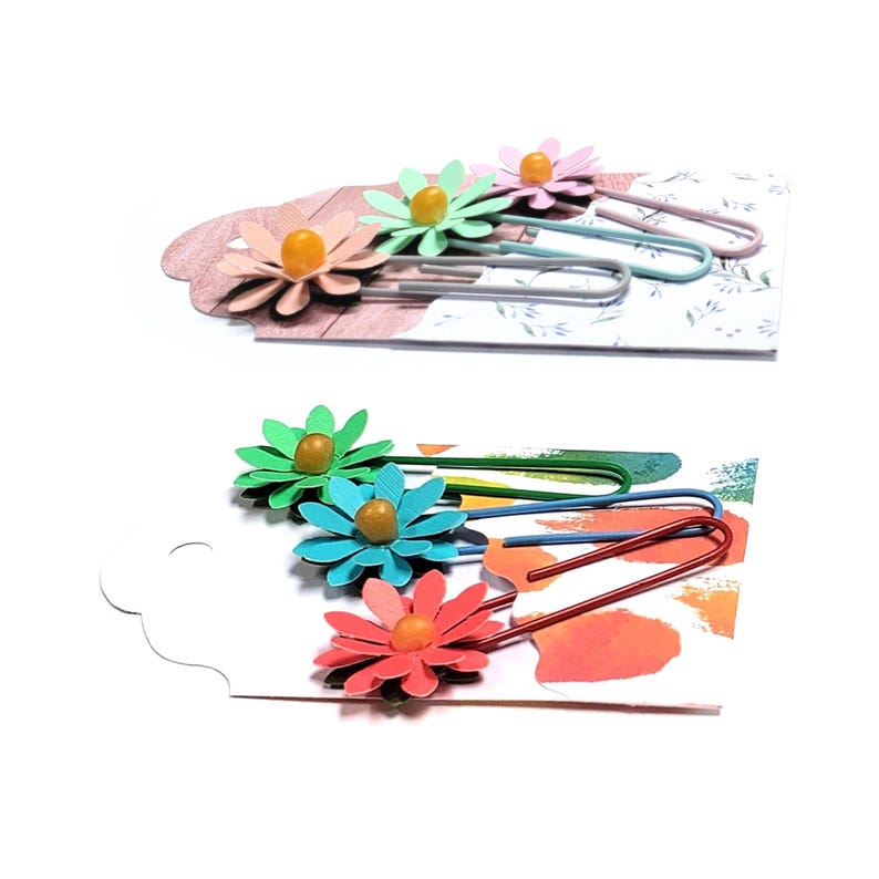 May include: Set of four colourful flower-shaped paper clips. Each clip has a yellow centre and a different coloured flower. The clips are on a white background.