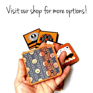 Op de afbeelding: Een hand houdt verschillende vierkante kaarten met Halloween-thema vast met oranje, zwarte en witte ontwerpen. De kaarten bevatten pompoenen, vleermuizen en andere griezelige afbeeldingen. De tekst op de afbeelding luidt: "Visit our shop for more options!"