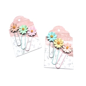 May include: Set of six colourful flower-shaped paper clips on a pink and white background. The paper clips are in shades of pink, blue, and yellow.