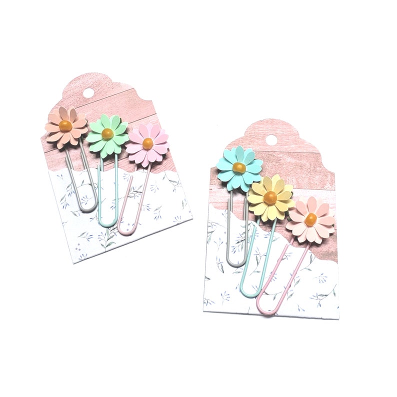 May include: Set of six colourful flower-shaped paper clips on a pink and white background. The paper clips are in shades of pink, blue, and yellow.