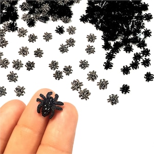 May include: A large quantity of black glitter spider embellishments. The spiders are scattered on a white surface. One spider is being held by a hand.