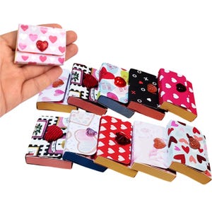 May include: A collection of small, decorative boxes with various designs, including hearts and floral patterns. The boxes are in shades of pink, red, and white, with some featuring glittery embellishments. The boxes are small and appear to be handmade.