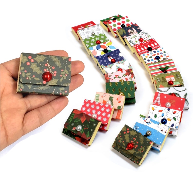 May include: Miniature Christmas-themed gift boxes in various patterns, including holly, poinsettias, and festive lights. Each box is square, with a flap closure and a decorative bead. The boxes are arranged on a white surface.